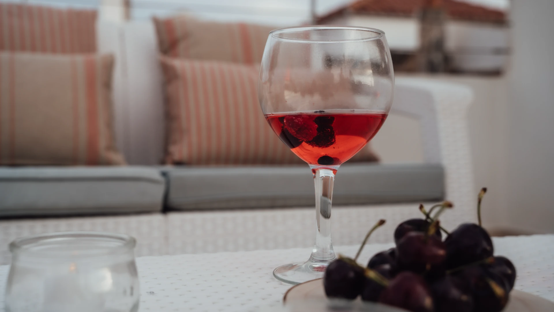 Copa de vino rosado con moras y cerezas en mesa frente a sof&aacute; con cojines.