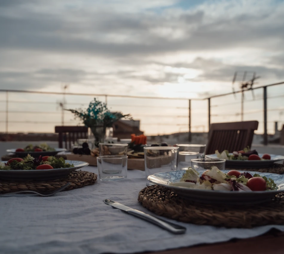 Mesa puesta al atardecer con platos de ensalada y vasos vac&iacute;os.