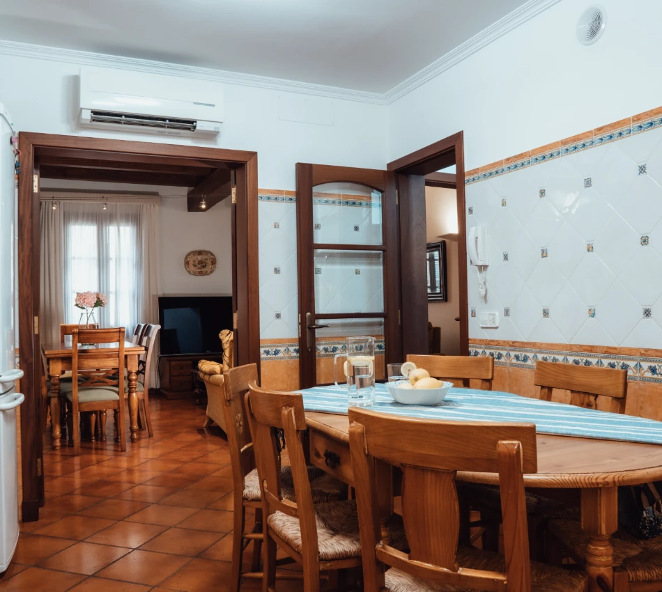 Comedor con mesa de madera, sillas y puerta abierta a sala con televisor.