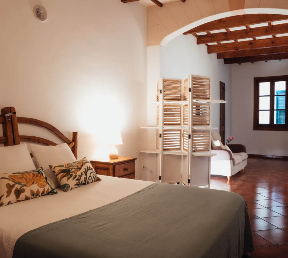 Habitaci&oacute;n acogedora con cama, l&aacute;mpara de mesa y sof&aacute; en fondo.