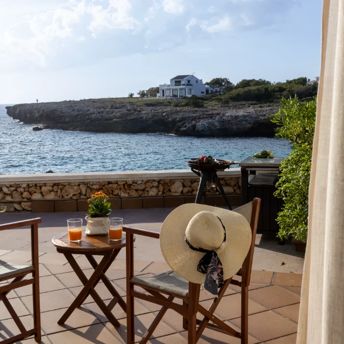 Terraza junto al mar con bebidas, sombrero y vista a una casa en el acantilado.