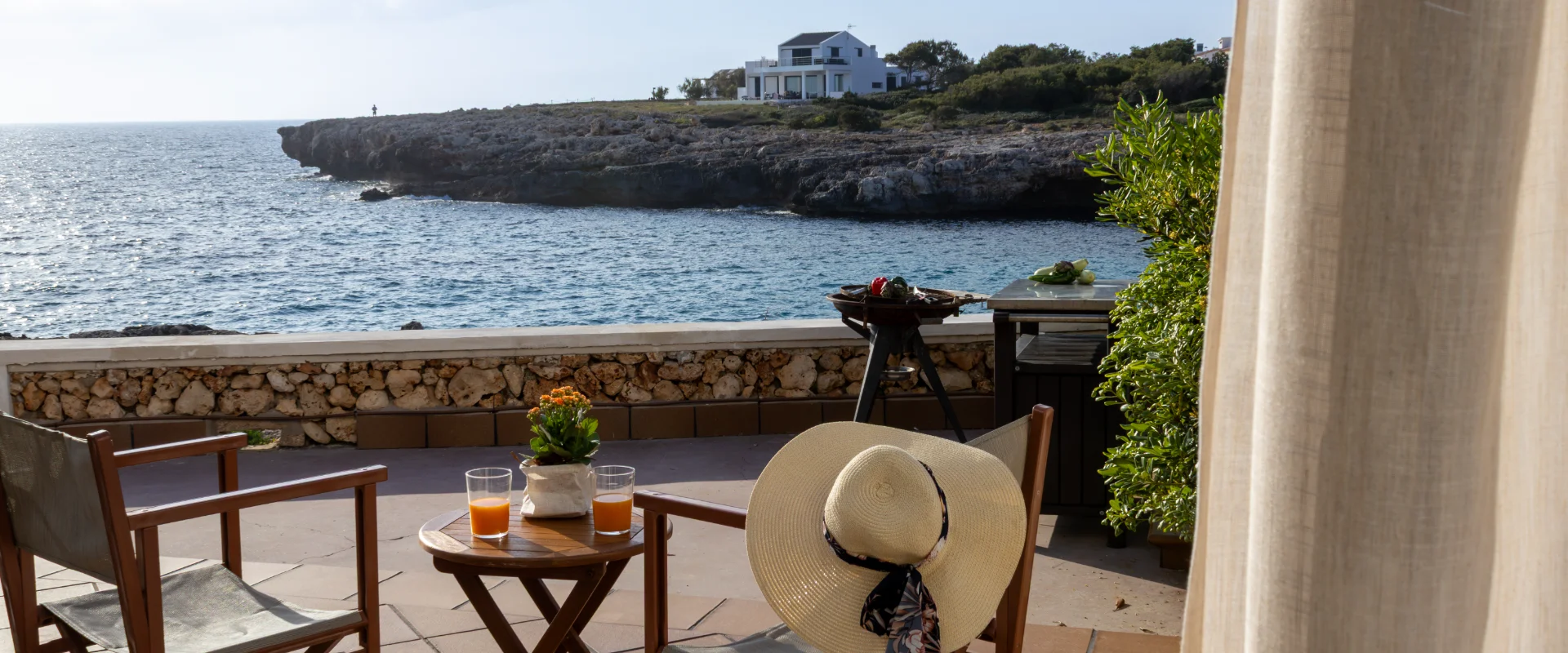 Terraza junto al mar con sillas, jugos y sombrero, vista a la costa.