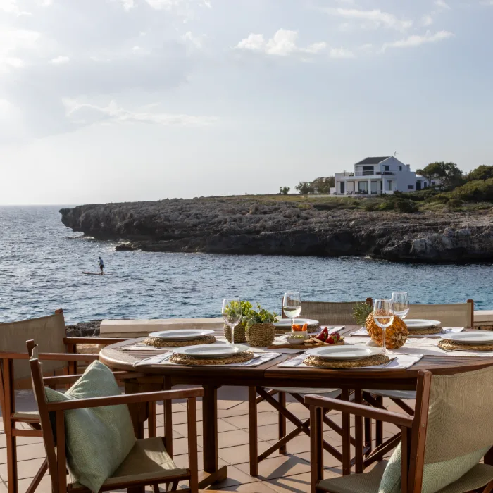 Mesa con platos y copas frente al mar, casa al fondo sobre el acantilado.