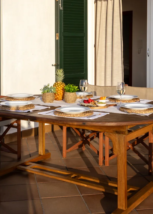 Mesa de madera en terraza con platos, copas y un centro de pi&ntilde;as y plantas.