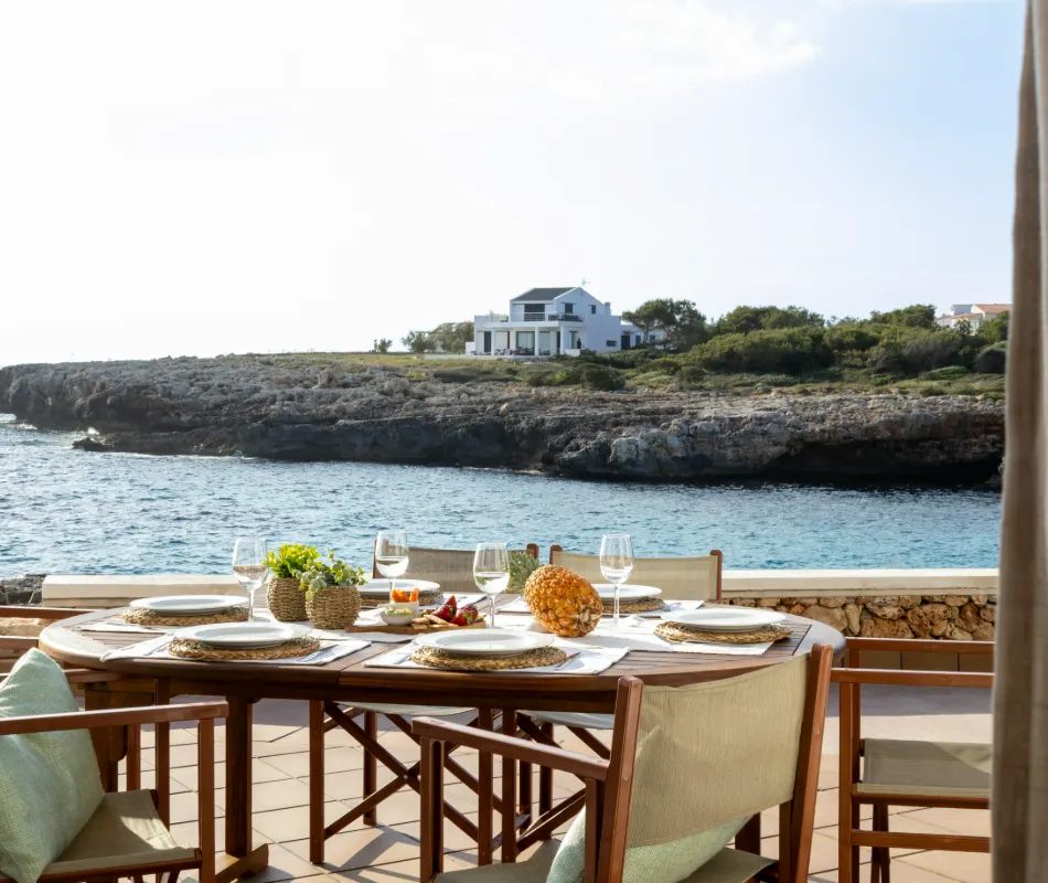 Mesa al aire libre con vista al mar y casa en la costa rocosa de fondo.