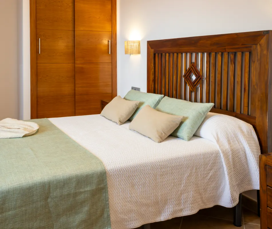 Cama con cabecero de madera y cojines en habitaci&oacute;n luminosa.