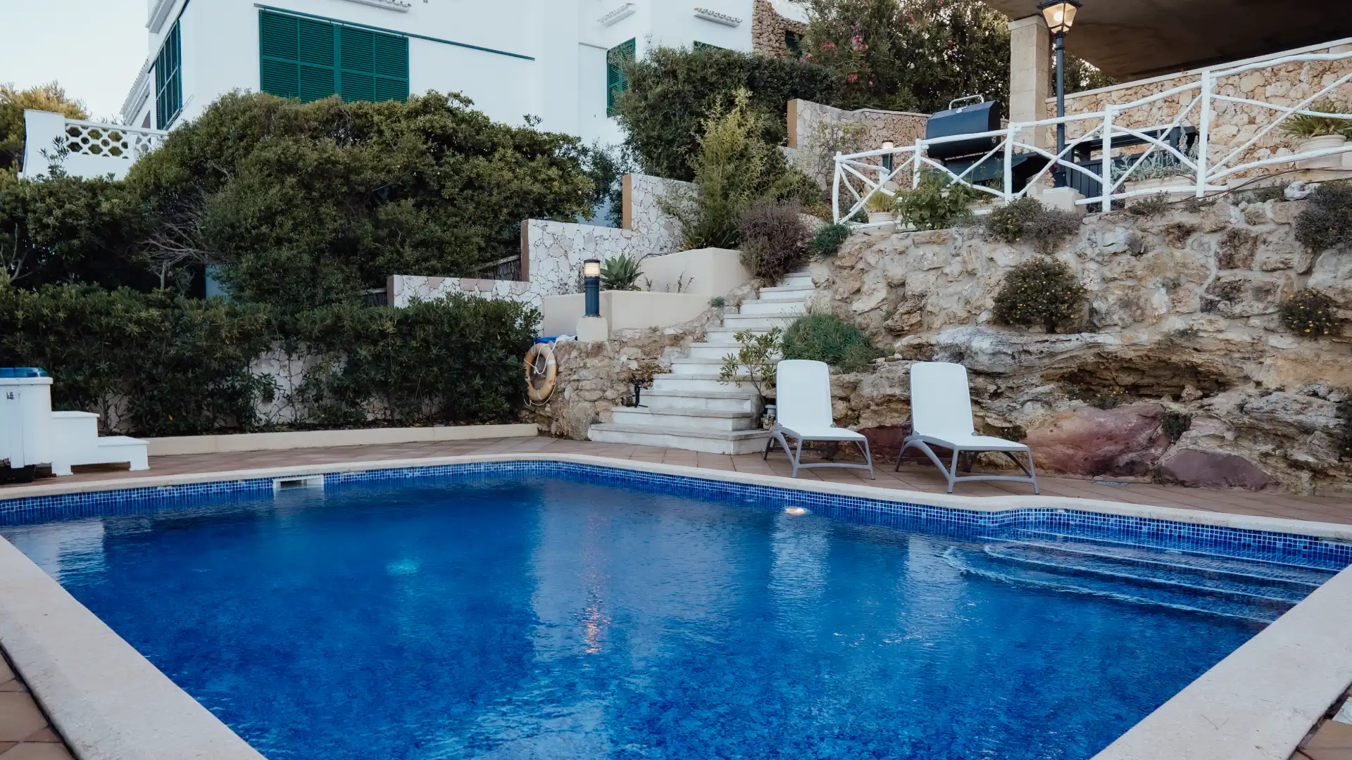 Piscina junto a escaleras de piedra y dos tumbonas blancas.