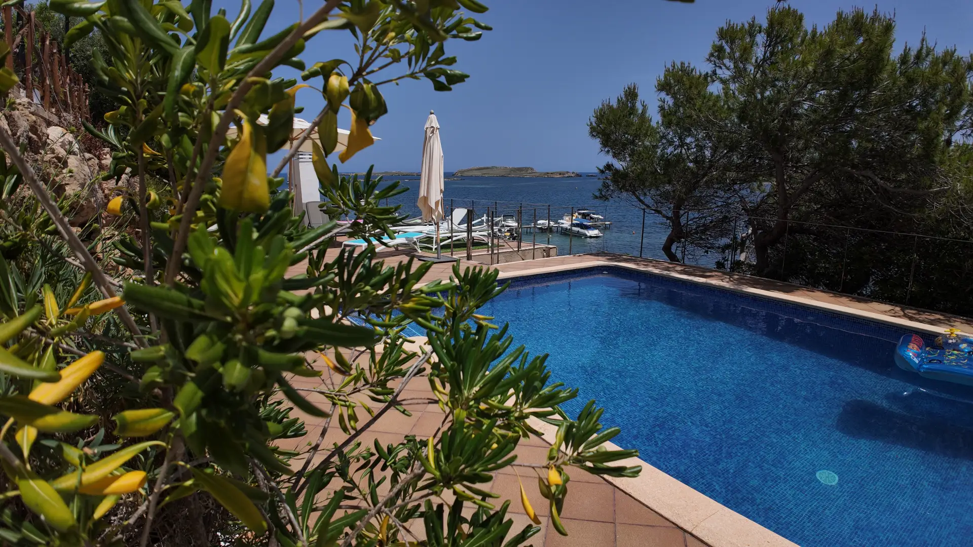 Piscina junto al mar, rodeada de &aacute;rboles y plantas bajo un cielo azul.