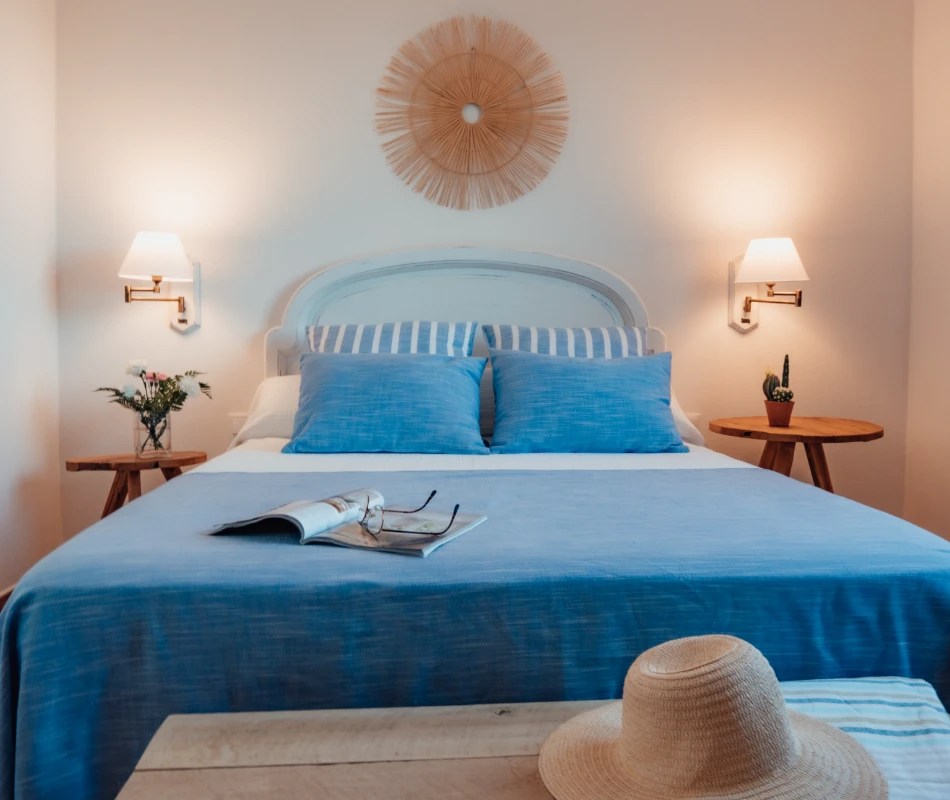 Cama azul con almohadas, l&aacute;mparas de pared y mesas con decoraci&oacute;n simple.