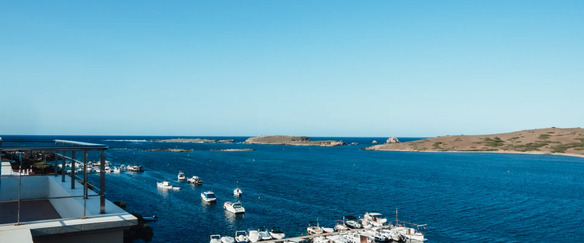 Barcos en un puerto deportivo con vistas al mar y la costa rocosa al fondo.