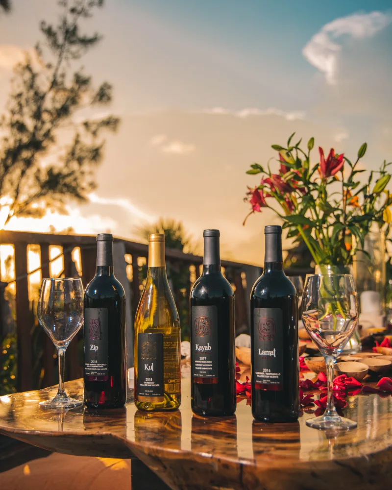 Four wine bottles on a table with glasses and flowers at sunset.