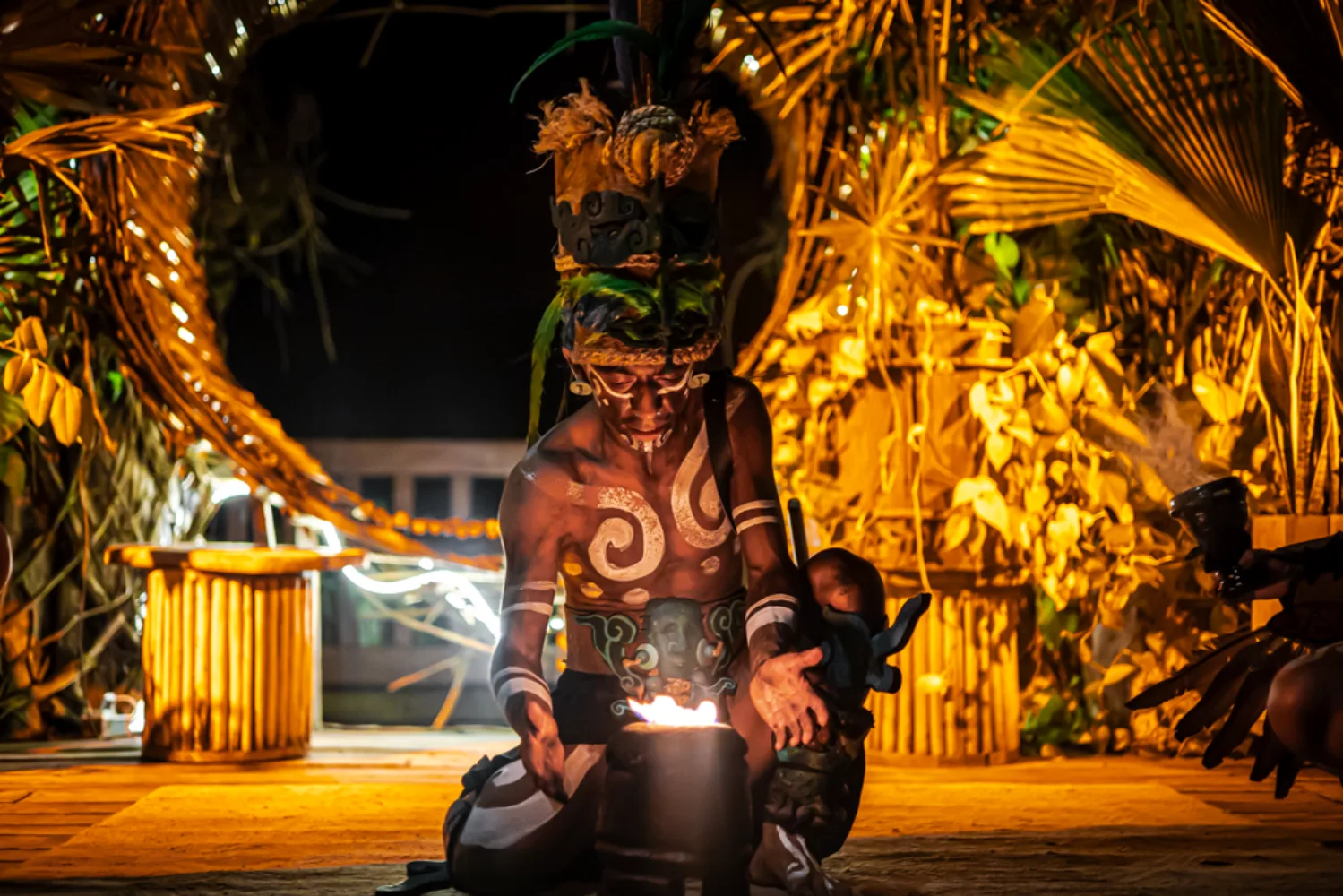 Person in traditional attire with face paint and fire, surrounded by foliage.