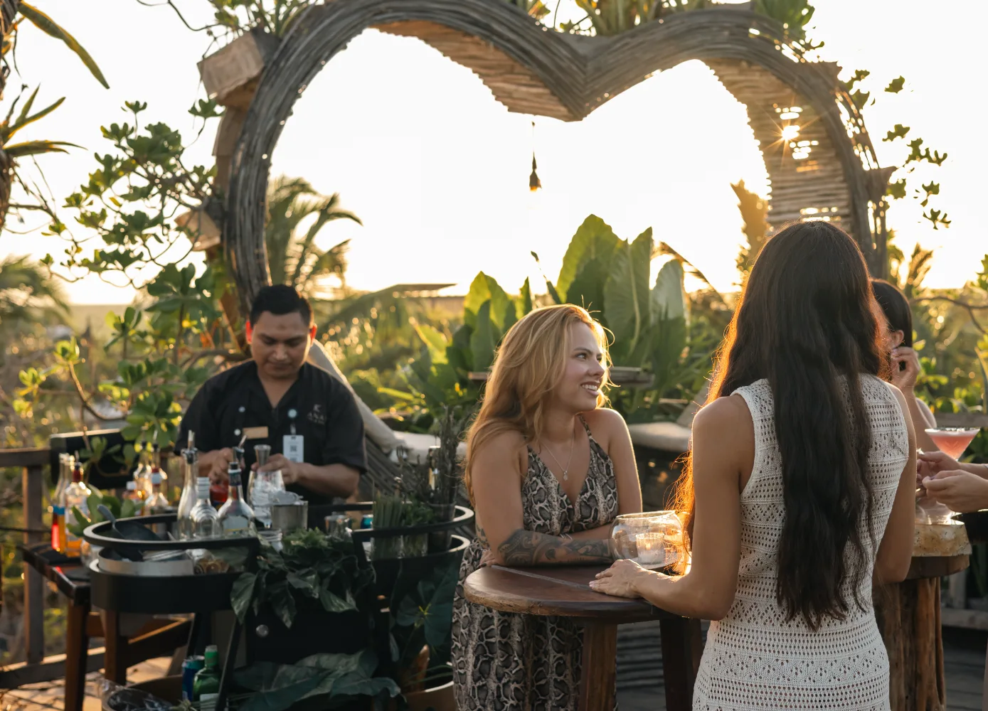 People enjoying drinks at a bar with a heart-shaped backdrop at sunset.