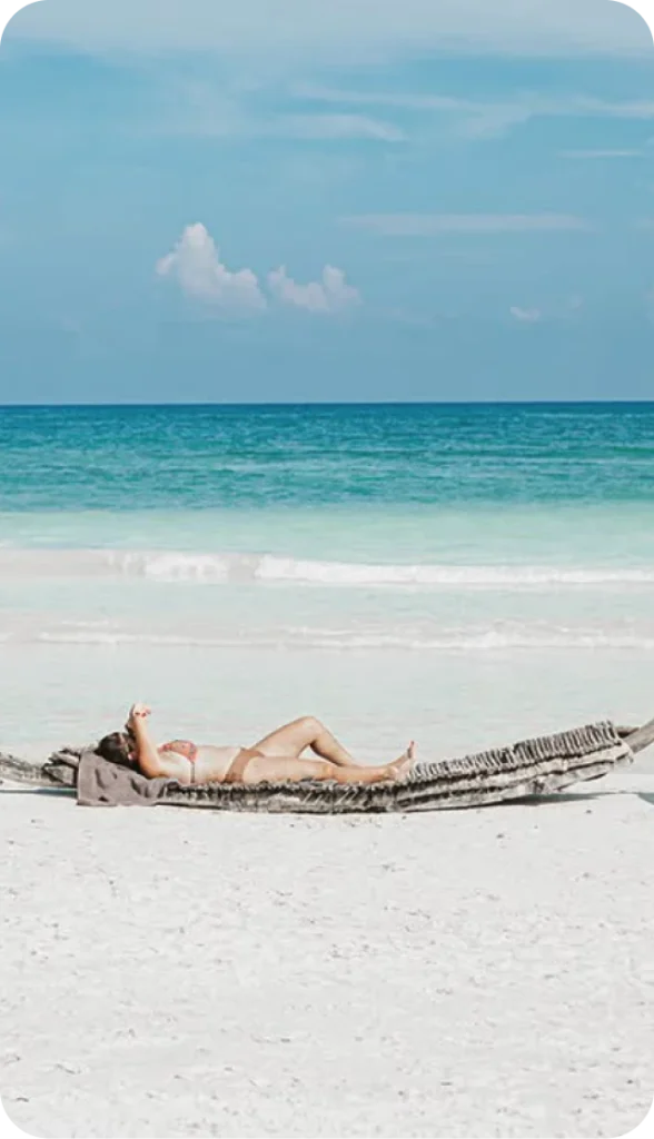 Person relaxing on a hammock at the beach with blue sky and ocean in background.