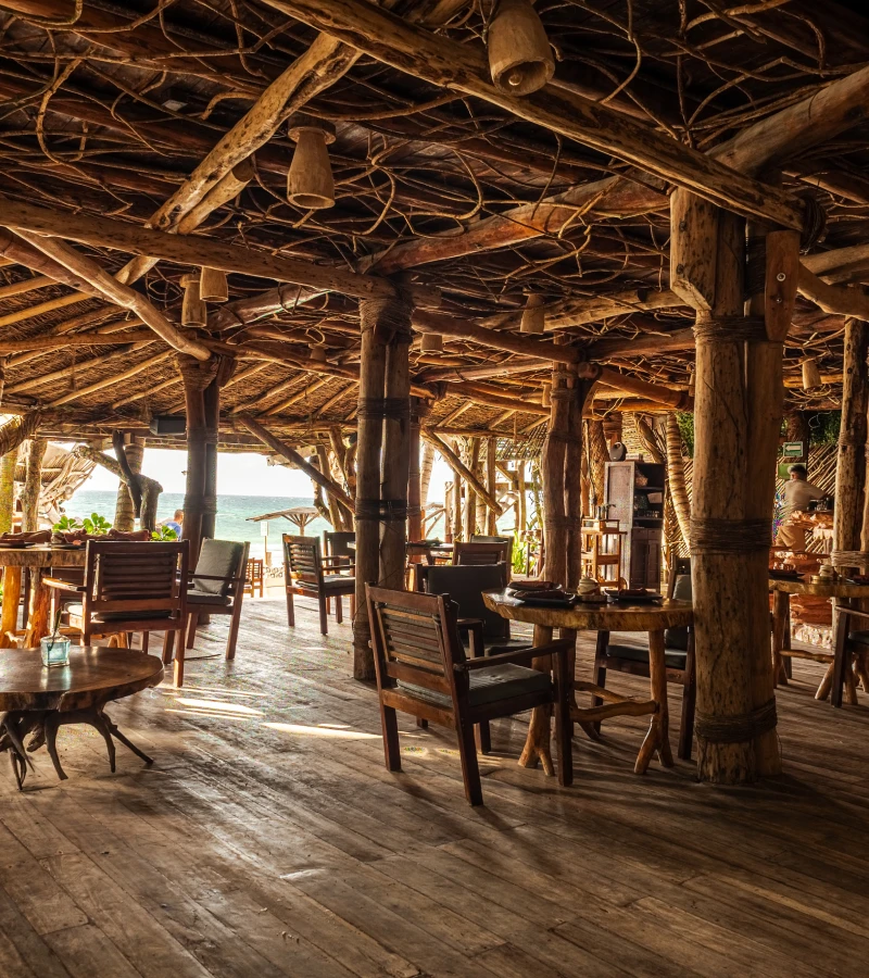 Rustic seaside restaurant with wooden chairs and tables, ocean view in background.