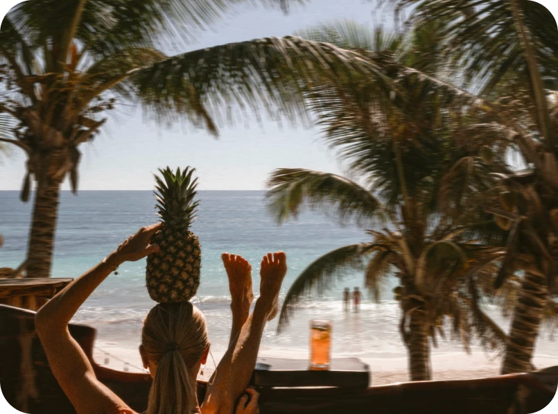 Person balancing a pineapple on their head at a tropical beach.