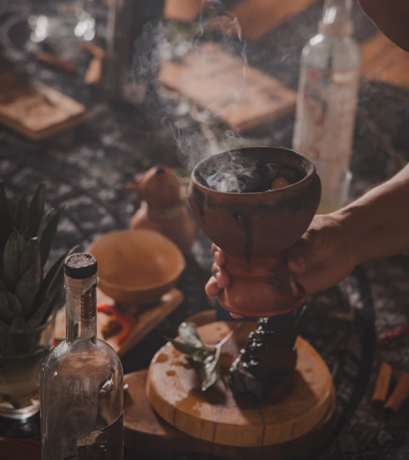 Hand holding a steaming clay pot, surrounded by bottles and leaves.