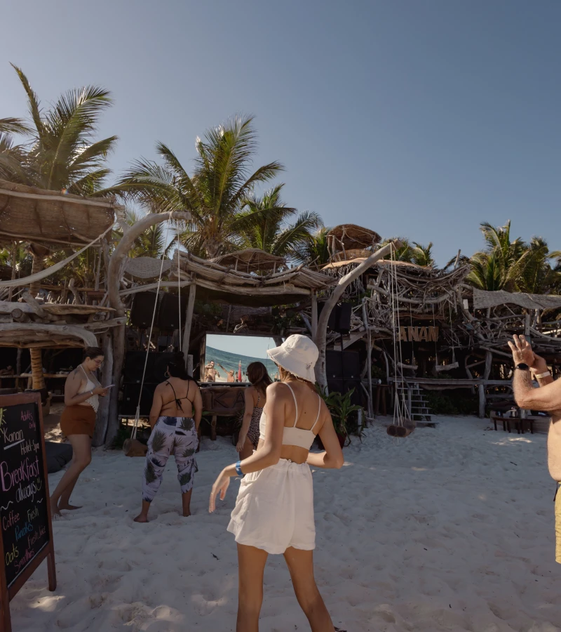 People at a beach bar with palm trees, wearing summer clothing and hats.