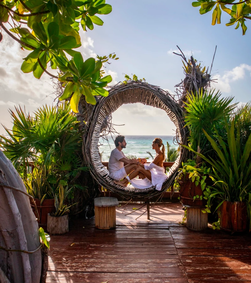 Couple sitting in a circular swing by the sea, surrounded by greenery.