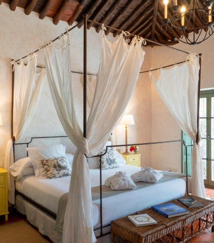 Cama con dosel blanco en habitaci&oacute;n r&uacute;stica con l&aacute;mpara de techo y mesas amarillas.