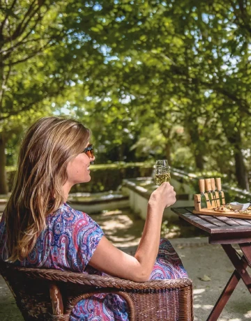 Mujer sentada en silla de mimbre al aire libre, sosteniendo una copa de vino.