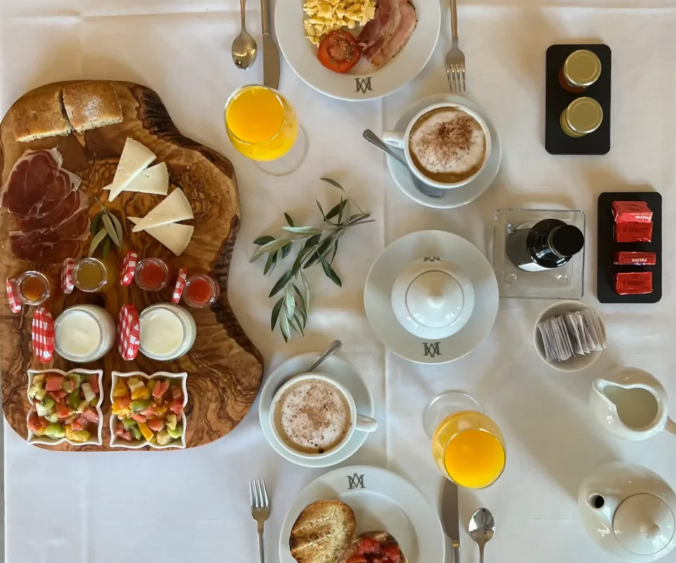 Desayuno gourmet con caf&eacute;, jugo, panes, embutidos y queso sobre mesa blanca.
