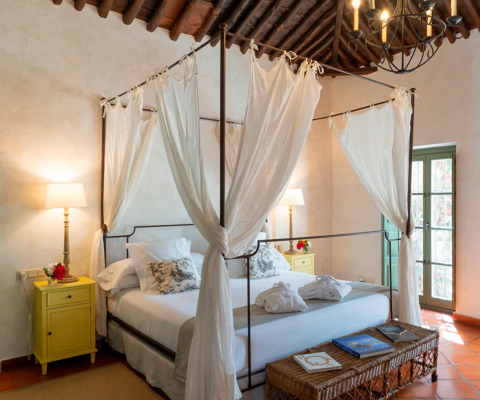 Dormitorio con dosel, mesita amarilla y ventanales al fondo.