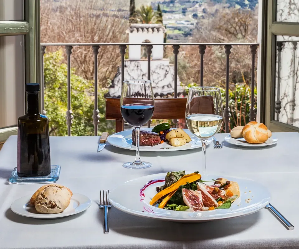 Mesa con platos de comida y copas de vino, vista a un jard&iacute;n desde el balc&oacute;n.