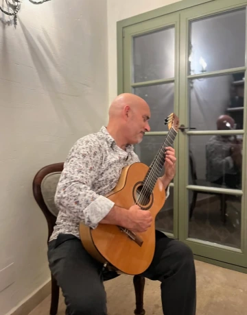 Persona tocando la guitarra ac&uacute;stica en una silla junto a una puerta de vidrio.