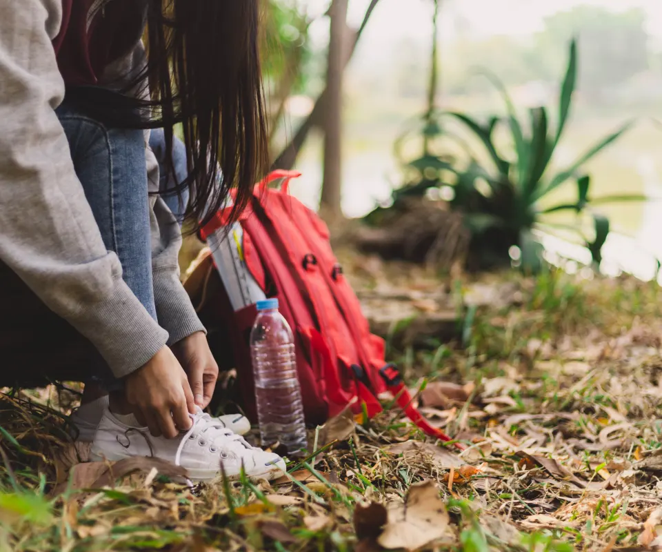 Persona atando sus zapatillas junto a una mochila roja y una botella en el campo.