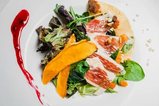 Ensalada gourmet con jam&oacute;n, mango y aderezo rojo en plato blanco.