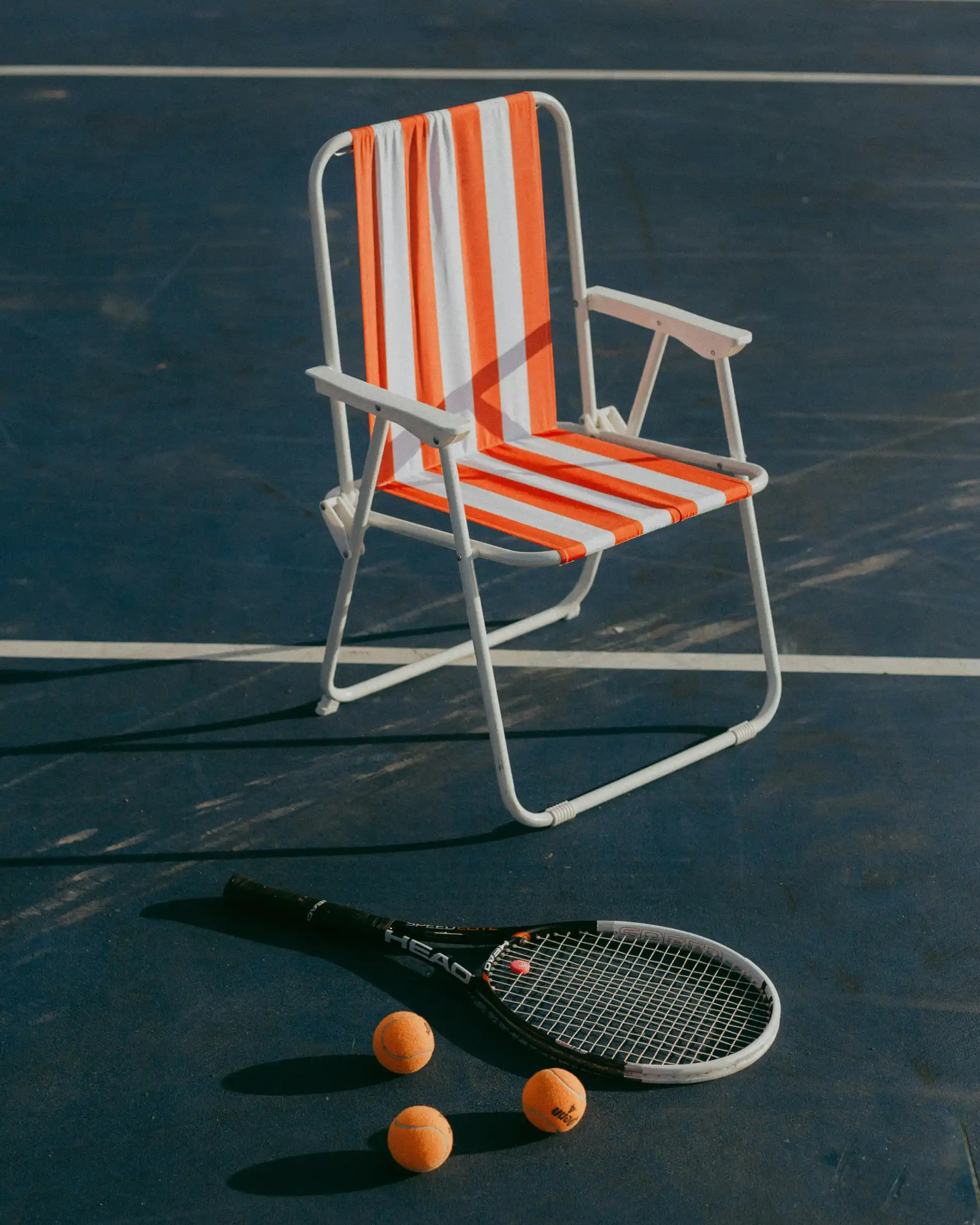 Chair with orange stripes, tennis racket, and balls on a court.