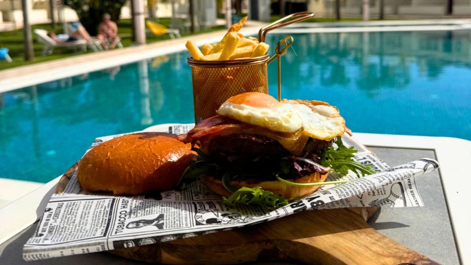 Burger and fries by a poolside with sunbathers in the background.