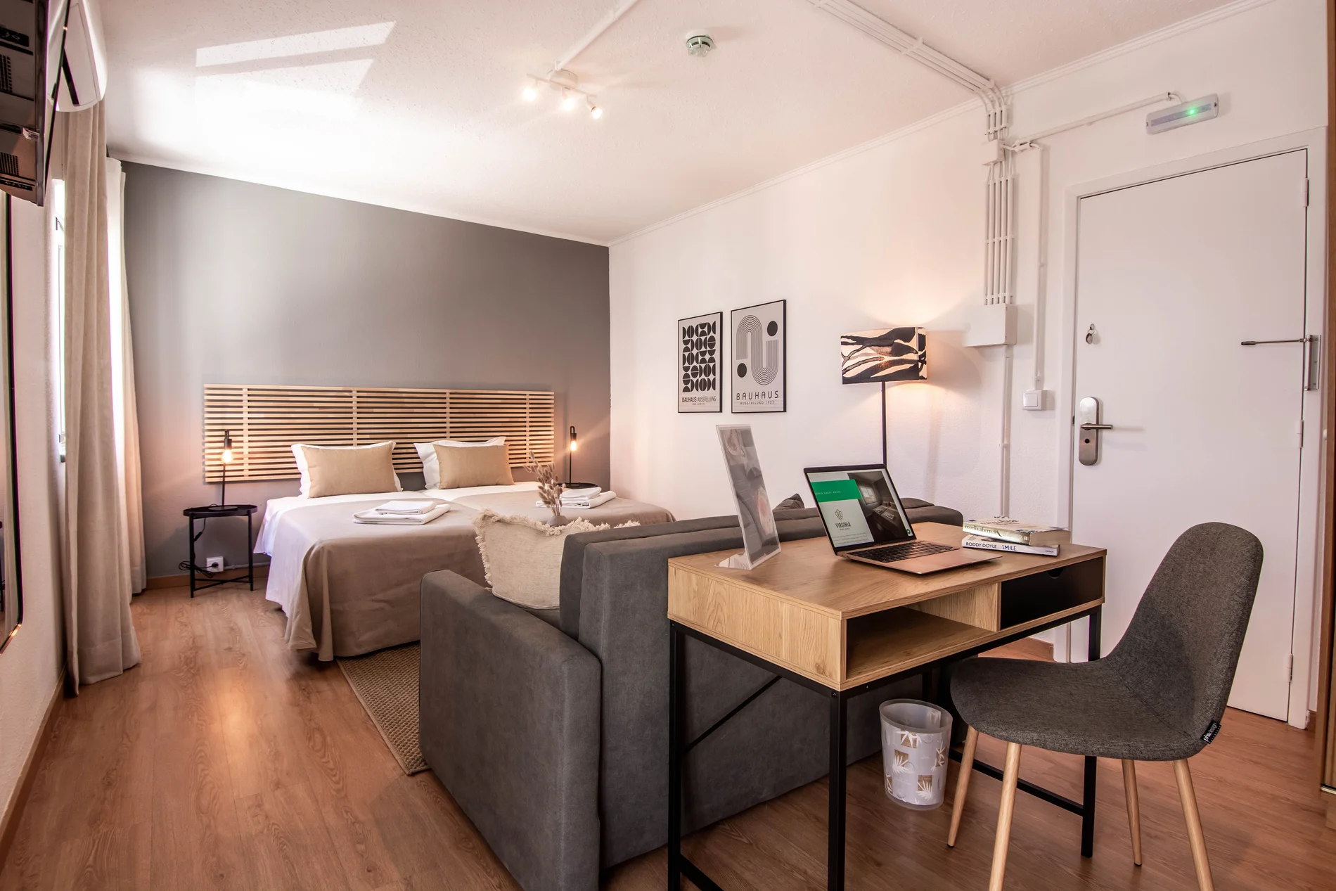 Modern bedroom with bed, desk, laptop, and chair, featuring minimalist decor.