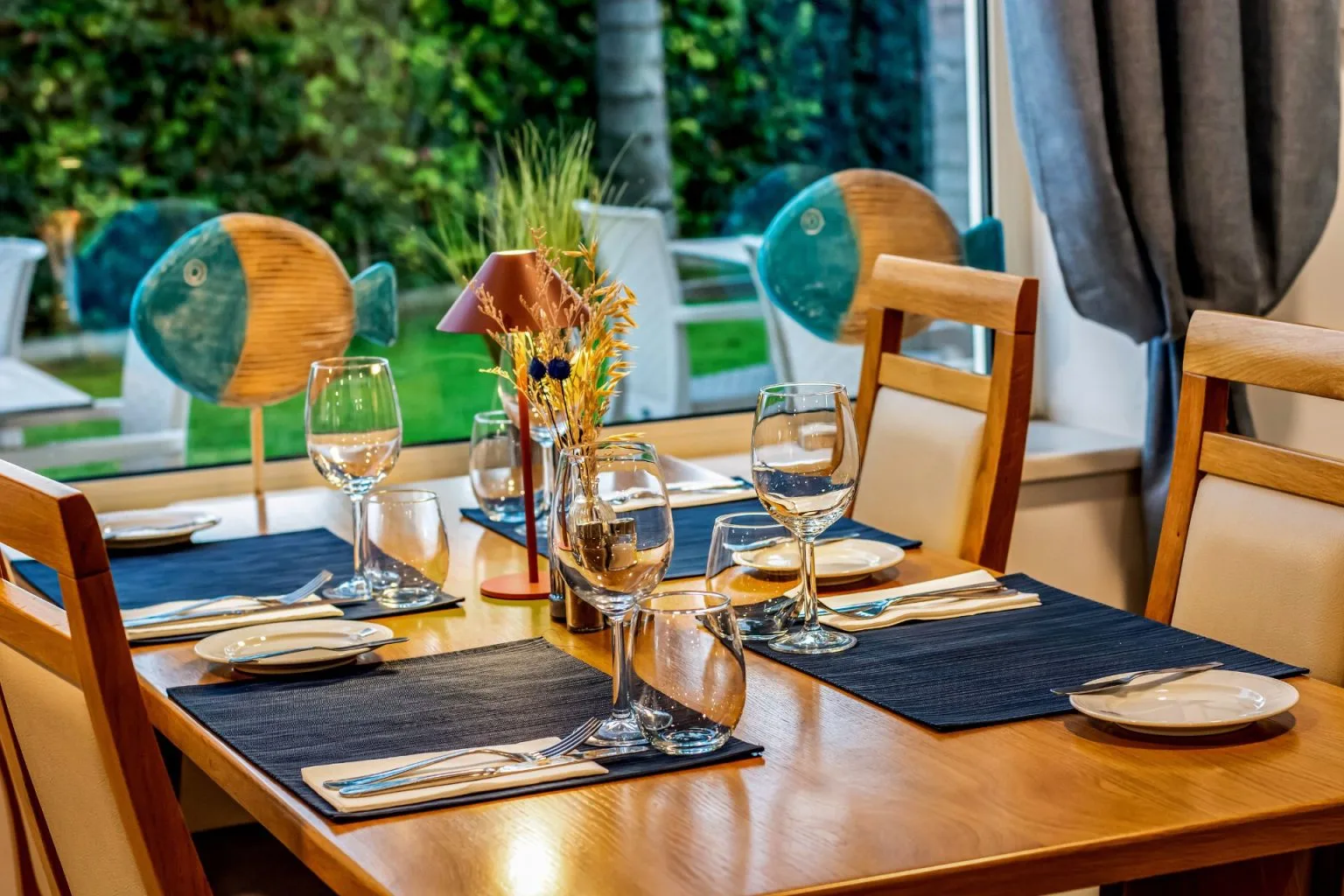 Dining table set for four with wine glasses and fish decor by a window.