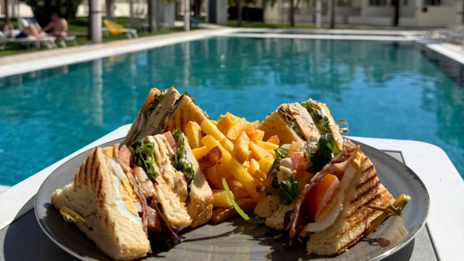 Club sandwich and fries on a plate by a poolside.