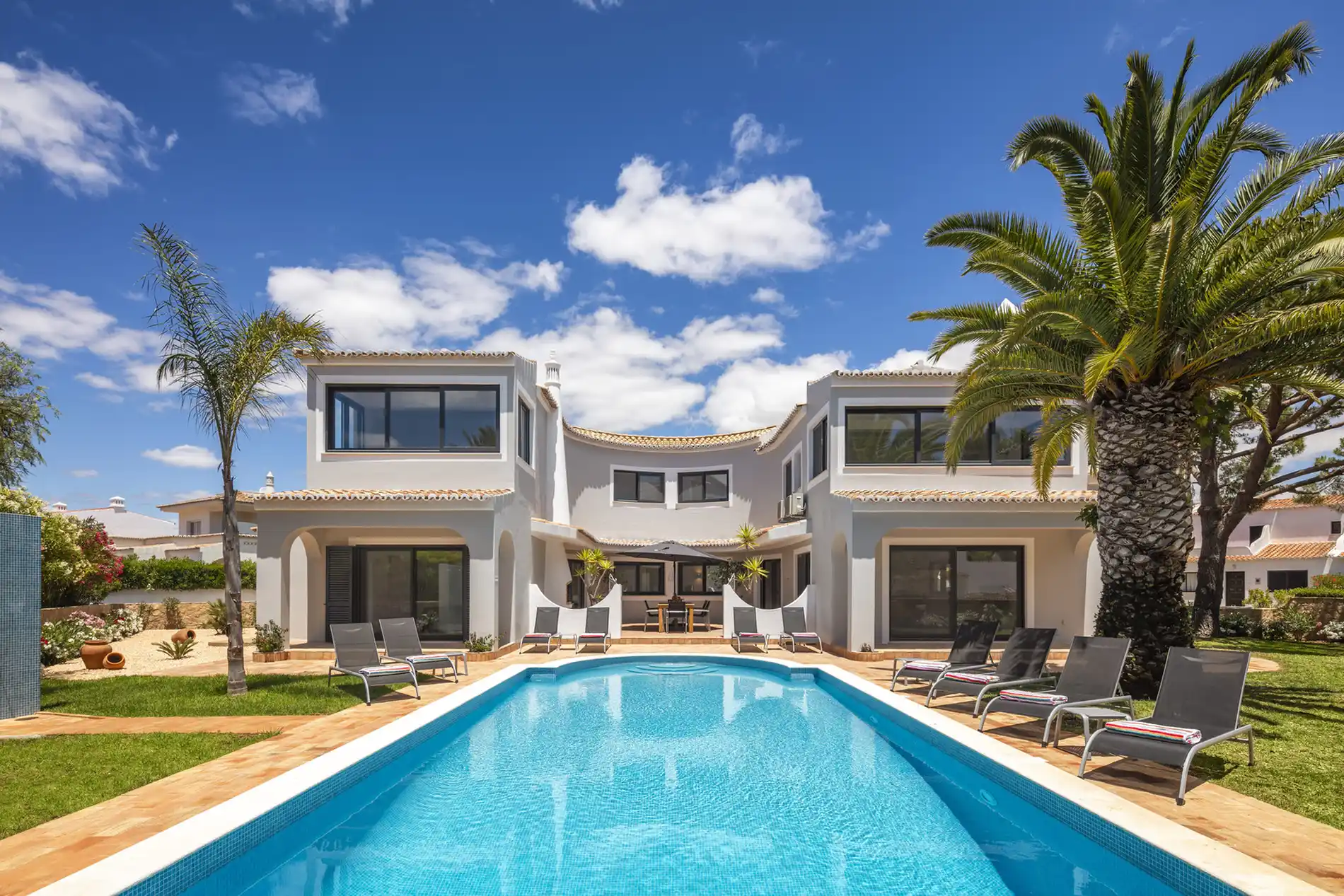 Modern villa with a pool, palm trees, and lounge chairs under a blue sky.