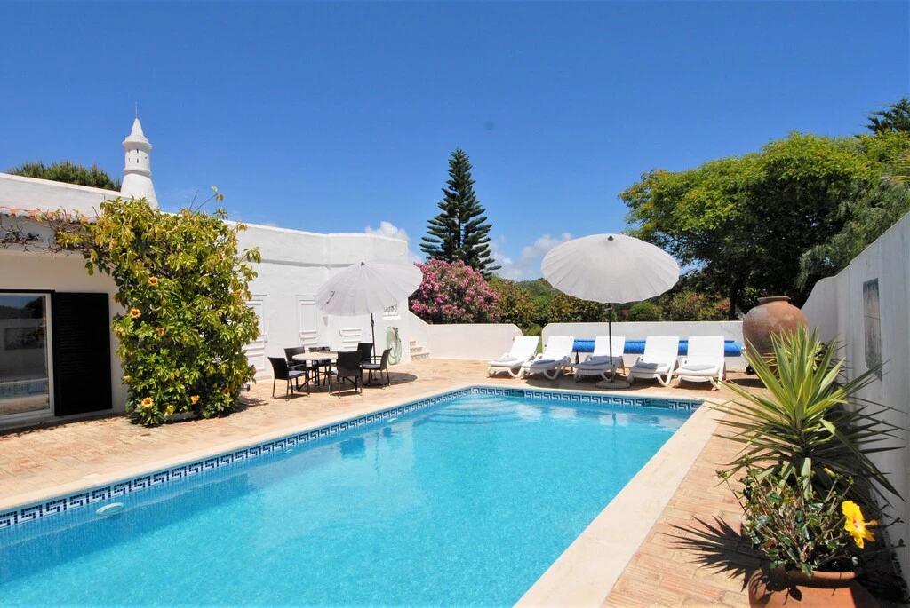 Sunny patio with a pool, lounge chairs, umbrellas, and lush greenery.