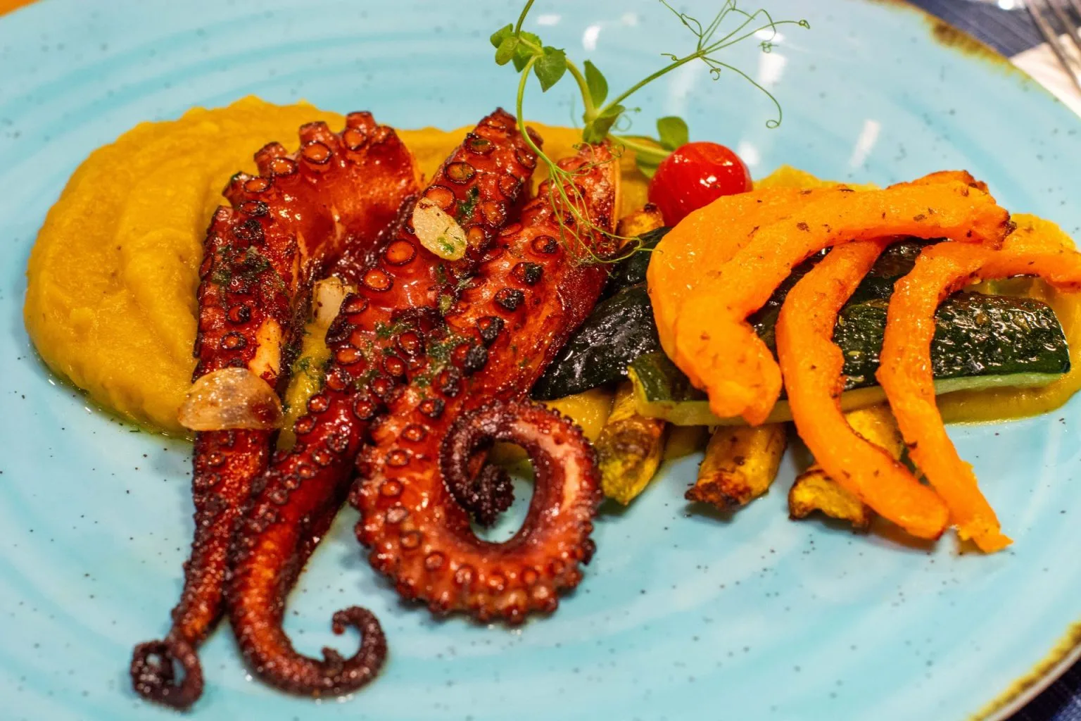 Grilled octopus tentacles with puree and vegetables on a blue plate.