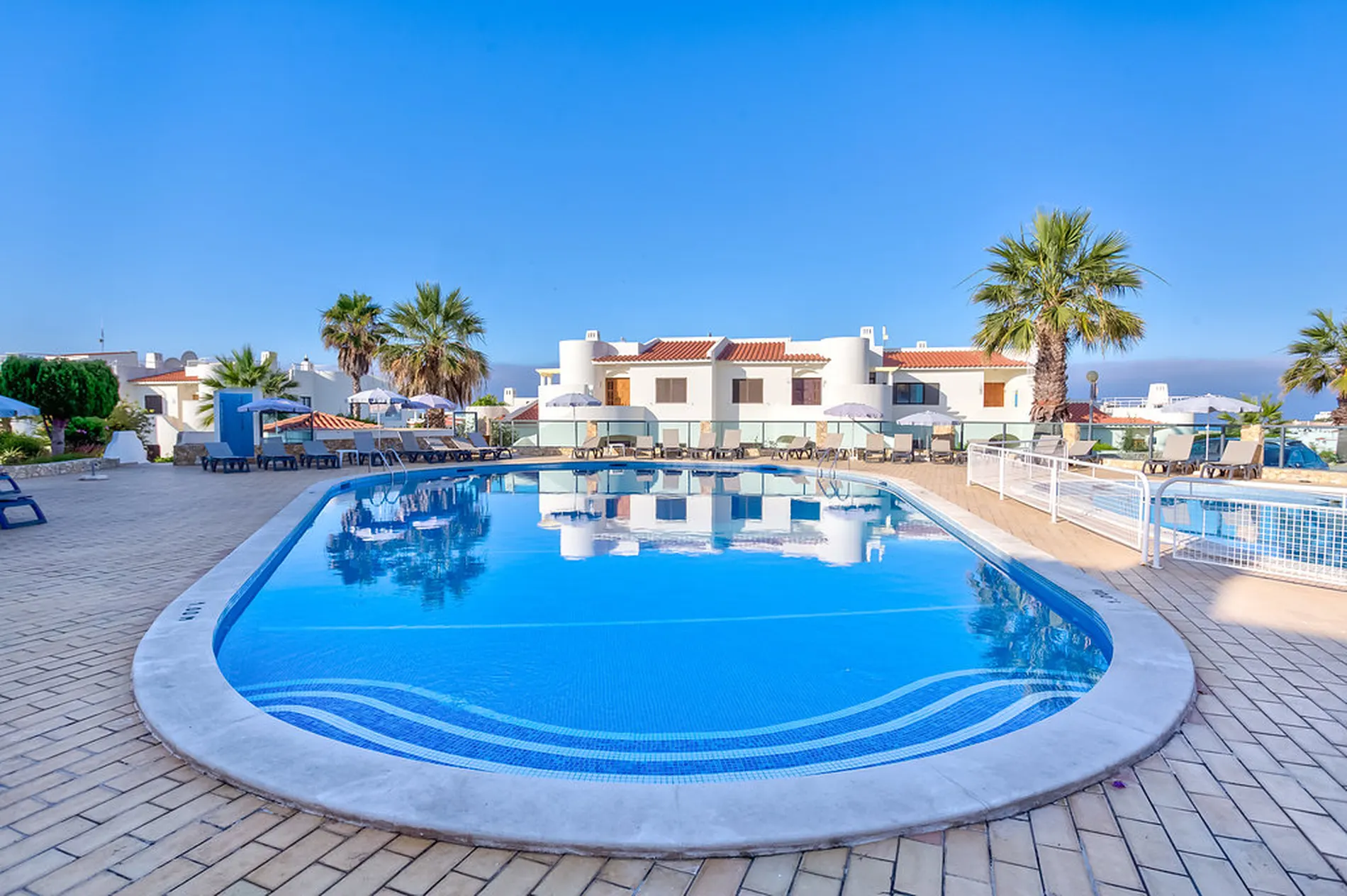 Poolside with palm trees and white buildings under a clear blue sky.