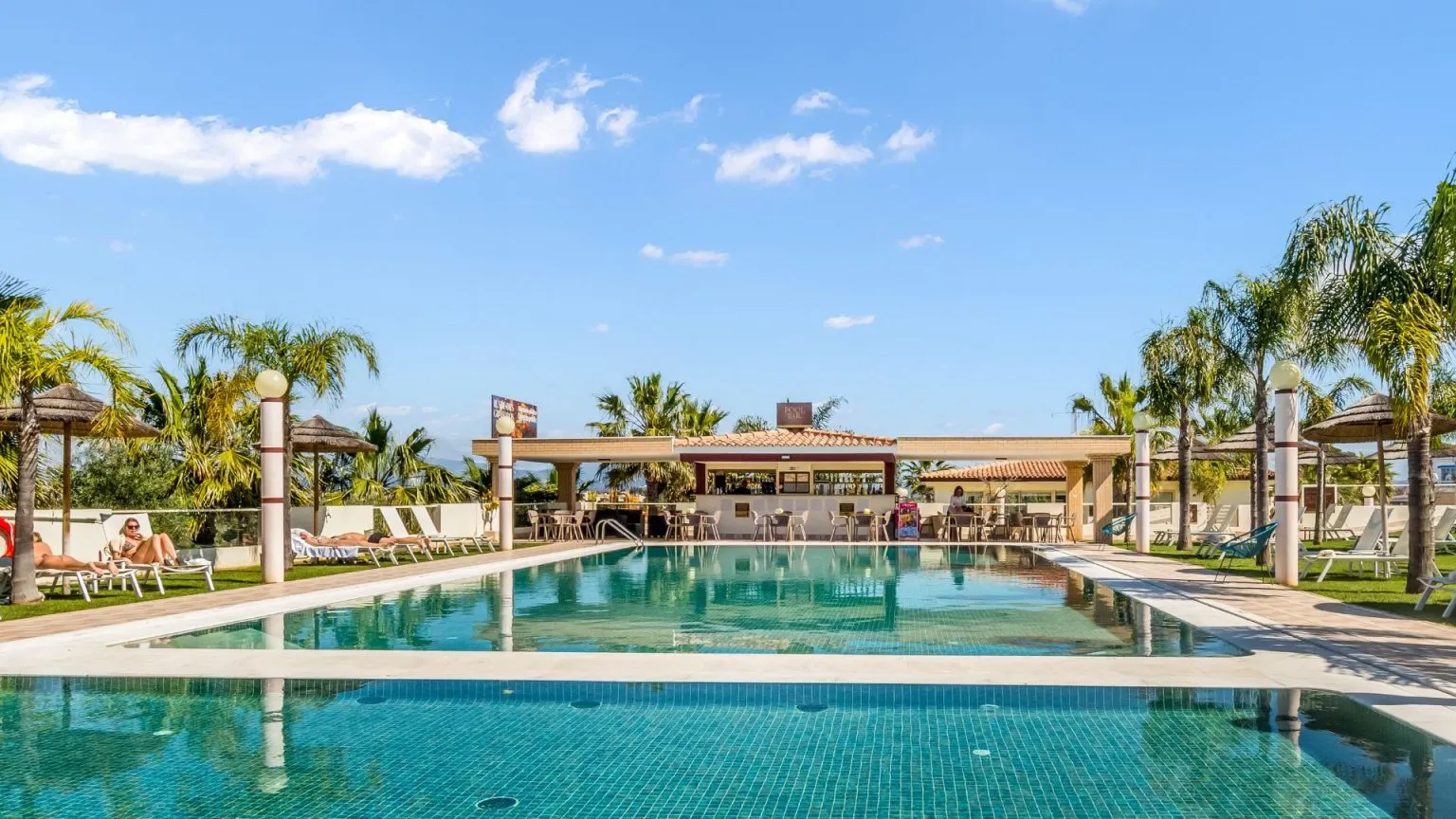 Outdoor pool with sun loungers, palm trees, and a bar under a clear blue sky.
