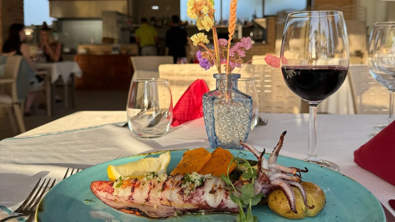 Grilled squid on a blue plate with wine, glasses, and flowers on a table.