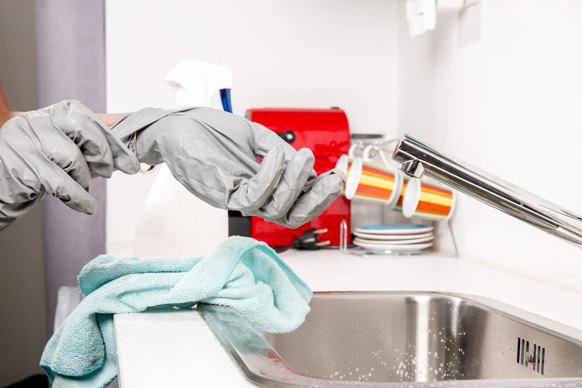 Hands in gray gloves near a sink with cleaning supplies and mugs.