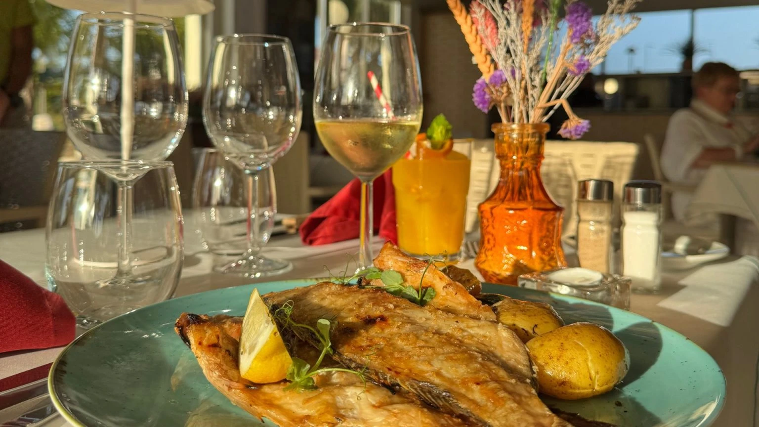 Grilled fish with lemon, wine glasses, and dried flowers on a table.