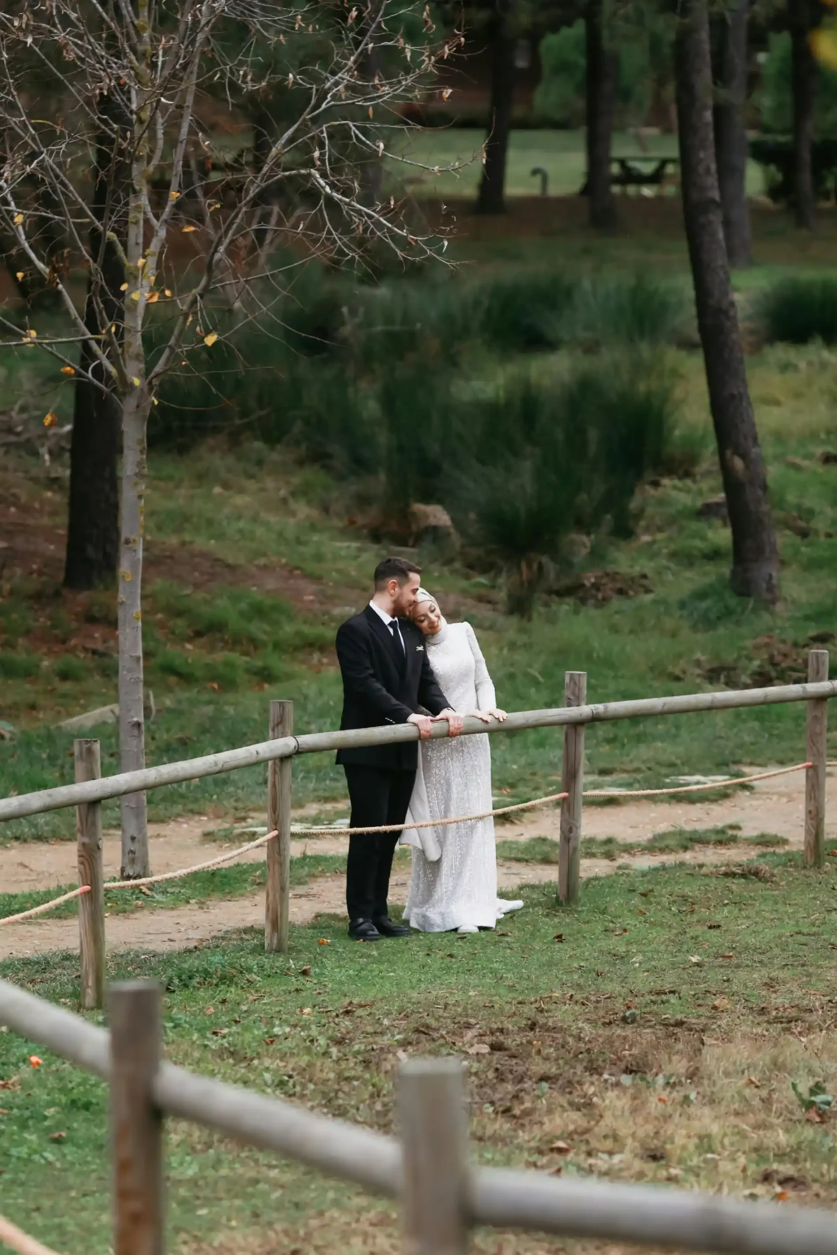 Bride and groom stand by a wooden fence in a serene, grassy park.