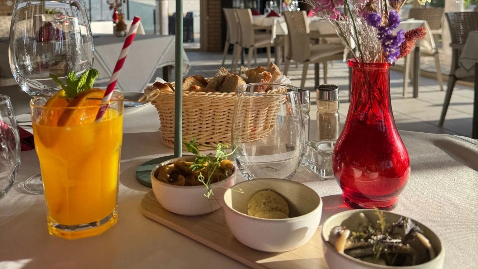 Glass of juice, bread basket, red vase, and bowls on a sunlit restaurant table.