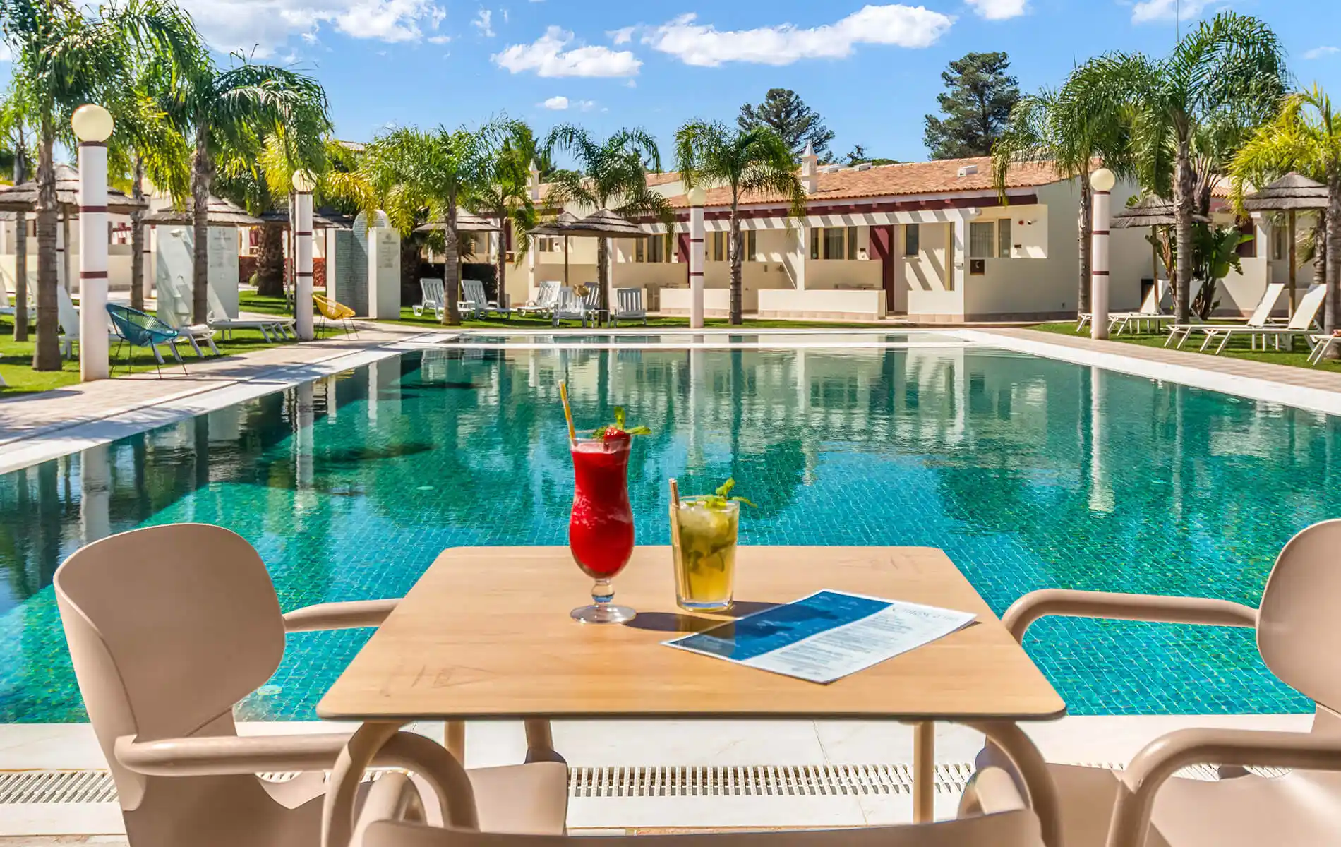 Poolside table with two drinks and a menu, overlooking a sunny resort pool.
