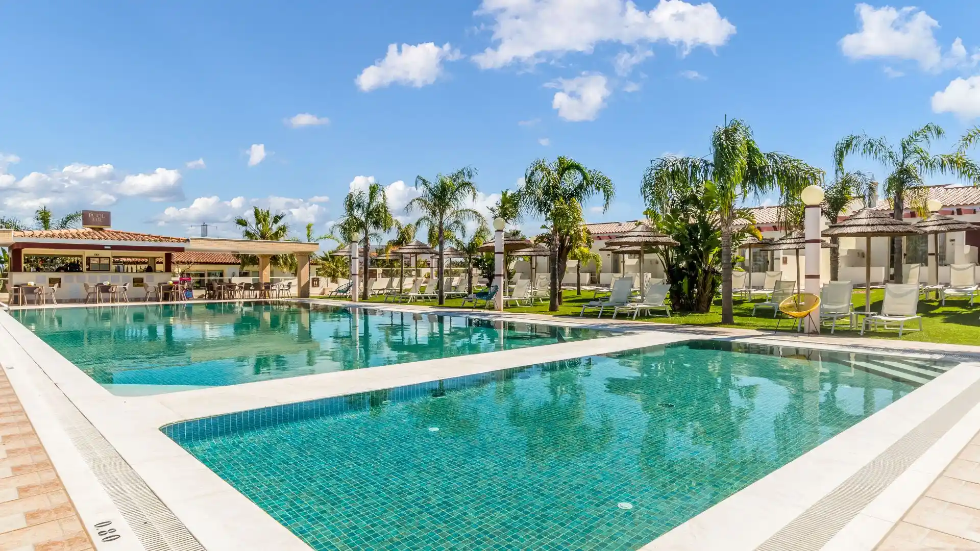 Outdoor pool with palm trees, sun loungers, and a poolside bar on a sunny day.