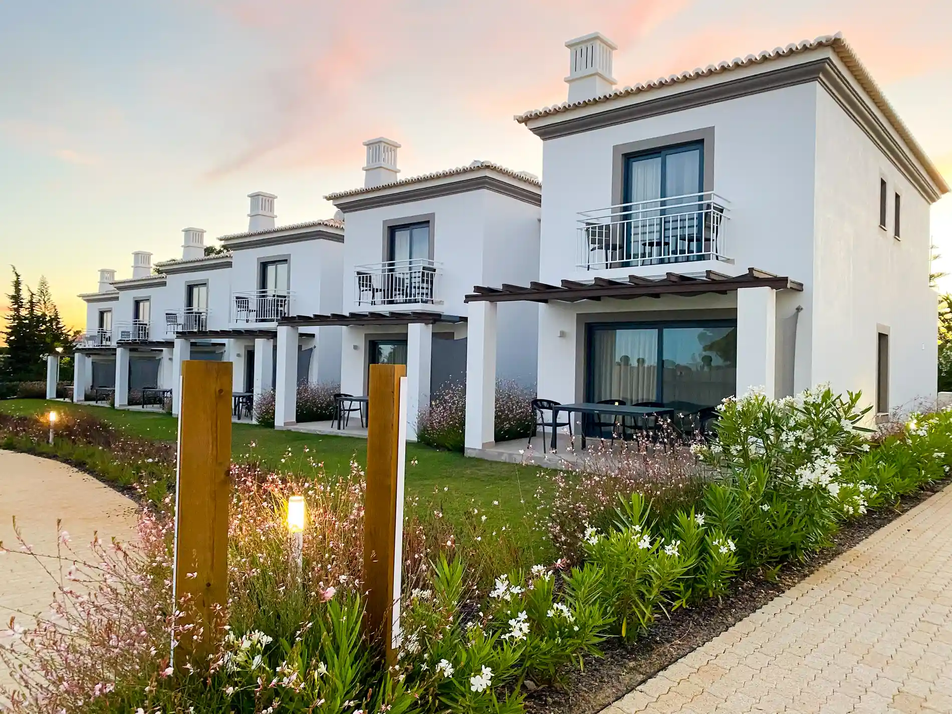 Row of white houses with balconies at sunset, surrounded by greenery.