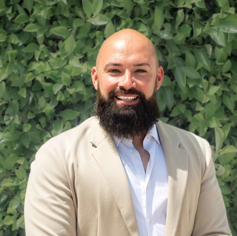 Man with a beard smiling in front of green leaves.